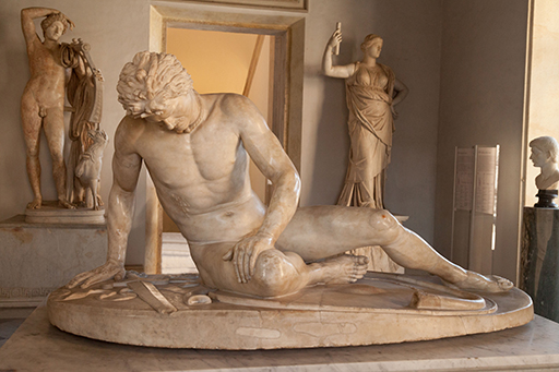 This photograph in landscape format shows the statue of a naked male figure collapsed in a seated position on an oval base, which is on top of a marble plinth. The figure leans to the left, supported by his right arm and looks downwards so that it is difficult to see his facial features; however, it is apparent that he has thick hair and a moustache and wears a torque. His right thigh rests on the ground with the right knee bent and the right foot under his left thigh; his left leg is extended to the side. His left hand rests on his right thigh. There is a vertical wound across his right ribs. A sword lies on the ground next to his right hand. The statue is in a gallery space, lit by light from a window to the right. Behind the statue is an open doorway with standing classical statues on either side of it.