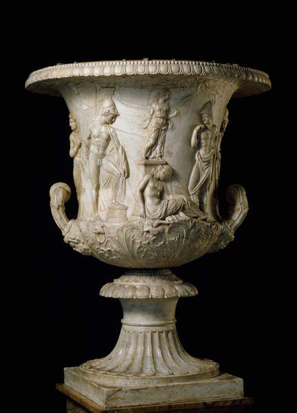 This photograph shows one side of a marble bell-shaped urn, lit from front left against a black background. A round fluted pedestal rises from a square base to support the inverted bell-shape, which has a fluted handle on either side and a raised acanthus leaf design around its base; a frieze represents a series of raised figures on the main body of the vase: from left to right: an athletic young man, facing right, stands with one foot raised on a block; then two young women are presented one above the other. The lower figure sits on the ground facing right, her head turned forward; her upper body is naked. The upper figure, smaller in scale, stands facing right in a diaphanous robe. The next figure is a partially naked and athletically proportioned young man facing left with his right arm bent so that his hand almost reaches his chin. A further figure is just visible on the left side of the urn.