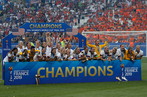 A photograph of the United States women’s national team standing behind a banner saying ‘Champions, France 2019’.
