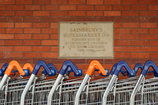 A photograph of shopping trolleys in front of a sign on a wall which reads ‘Sainsbury’s supermarket Ltd. Former site of Scunthorpe United’s old show ground 1899-1988.