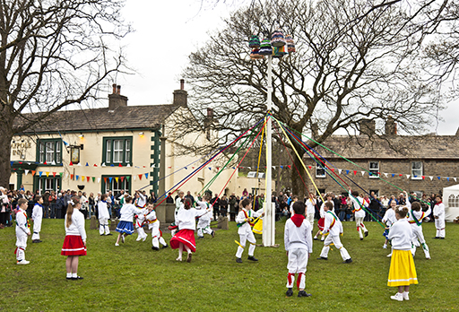 This is a colour photograph of a group of children dancing around a tradition Maypole on a village green.