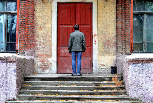 Man standing outside a house with a red door. The house is run down with flaking paint, stained windows and graffiti.