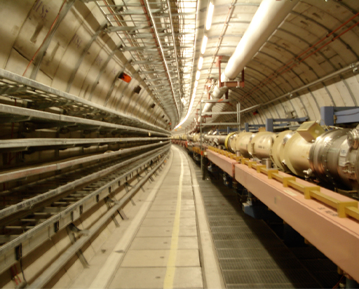 This is a photograph of the inside of a particle accelerator tunnel. It’s a rounded industrial setting stretching a long way into the background. Machinery and pipes run along the tunnel.