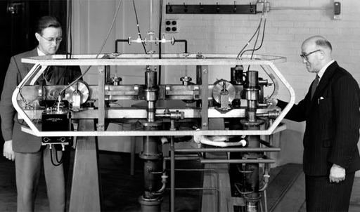 This is a black-and-white photograph of two men examining the first practical atomic clock.