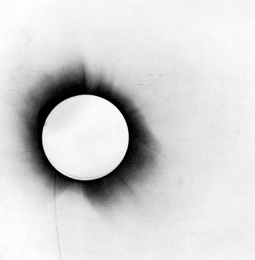 This is a photographic negative of the eclipse of 1919. The Moon is completely eclipsing the Sun. Sunlight around the edges is seen in black. The rest of the image is grey-toned. There are some thin horizontal lines seen to the upper right of the image.