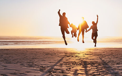 Four people including three children holding hands and jumping on the beach as the sun sets