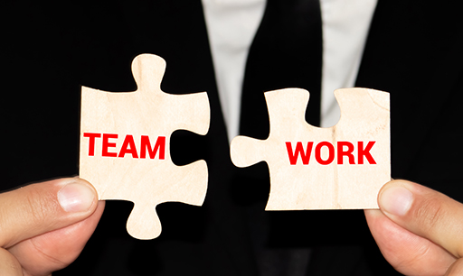 Two hands are holding jigsaw pieces. One has the word ‘team’ written on it, the other has the word ‘work’ written on it. The hands are pushing these pieces of the jigsaw together.