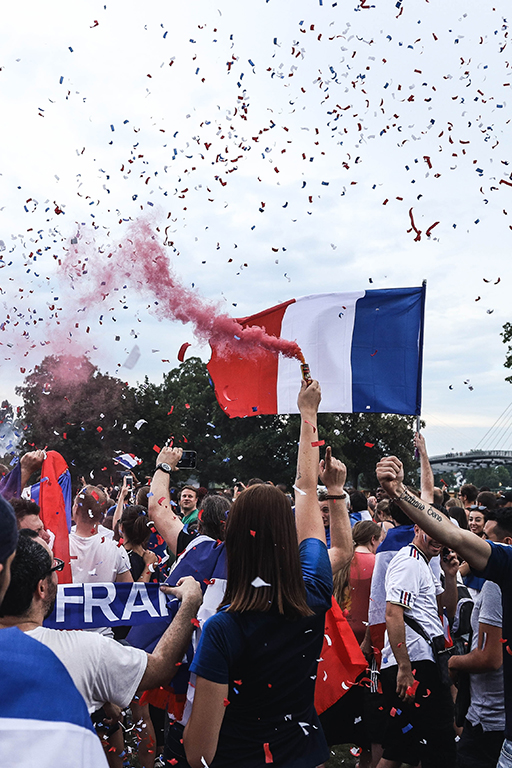 A photograph of a crowd with French flags and setting off flairs of the French flag’s colours.
