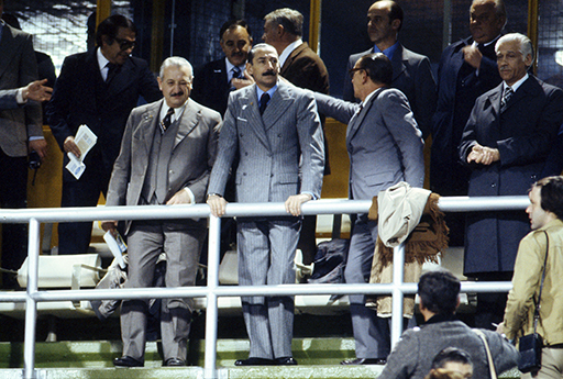 A photograph of a group of men wearing suits in a football ground.