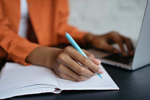 A photograph of a person writing in a notepad while on their laptop.