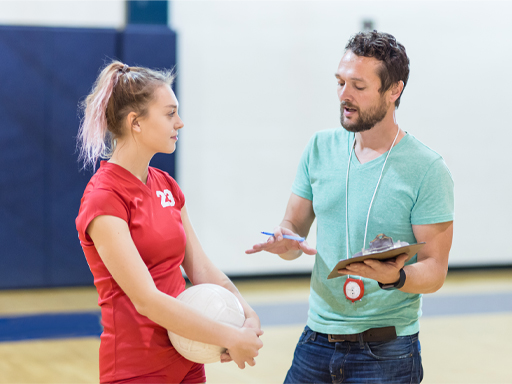 The image shows a male coach talking to a female athlete. She is holding a volleyball.