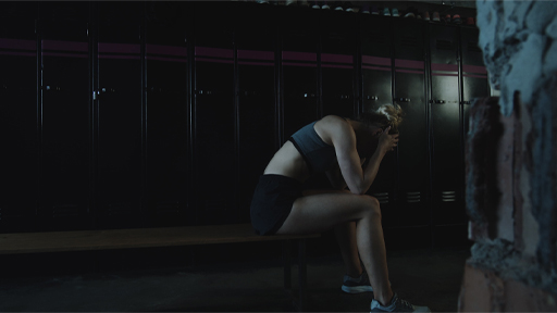 The image shows a female in exercise clothing sitting on an exercise bench in a darkened room. She has her head in her hands.