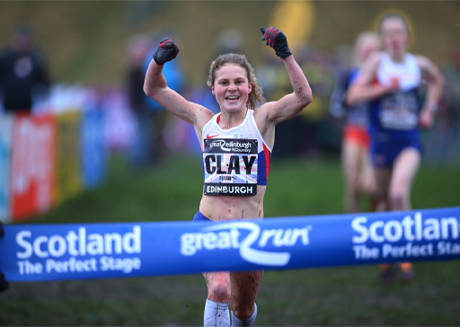The image shows the athlete Booby Clay winning a cross country running race. She is about to run through the finish tape and is celebrating by having her hands above her head and smiling.