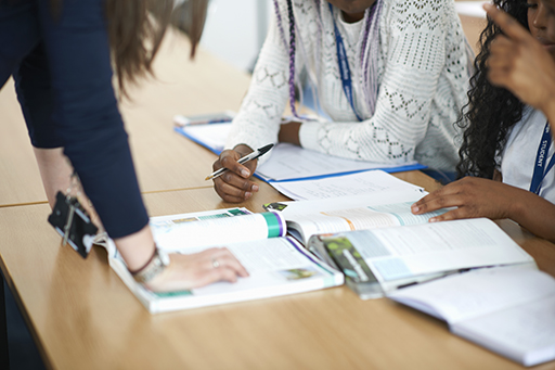 A photograph of two people at a desk working through a textbook. There is someone standing on the other side of the table talking to them.