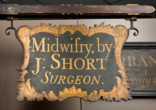 A photograph of a wooden shop sign with the text ‘Midwifry by J. Short Surgeon’ painted in gold. The background of the sign is painted dark, with the edges decorated in a gold pattern.