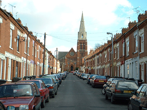 A colour photograph of a residential street. Cars are parked on both sides of the road. On either side of the photograph are rows of red-brick terraced houses. At the far end of the street there is a red-brick church with a tall spire. There are no people visible in the photograph. Streetlights and telephone wires are visible in the image.