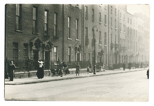 A black and white photograph of a street. There are no vehicles visible in the image, but there is a road and a pavement for pedestrians. Several people are visible in the image, including a nun and a woman with a pram. Behind them stand a row of brick houses, most with railings.