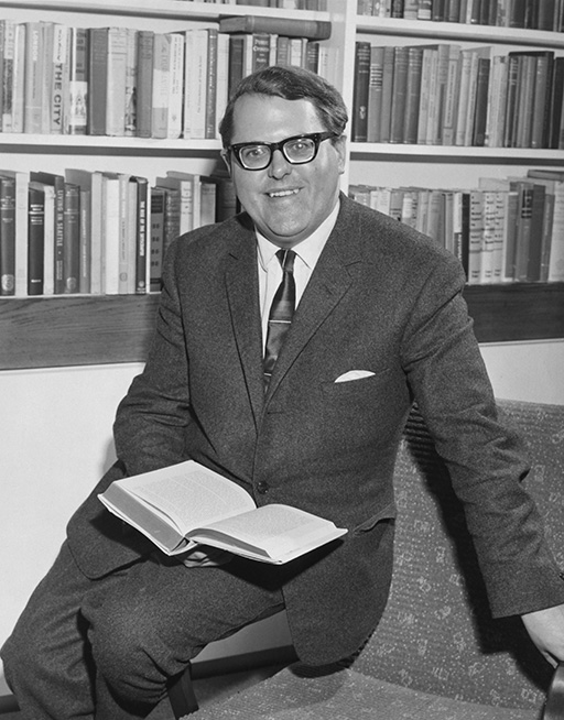 A black and white photograph of a smiling man. The man wears a suit, tie, and glasses. He sits on the arm of a chair in front of a bookcase and holds an open book in his lap.