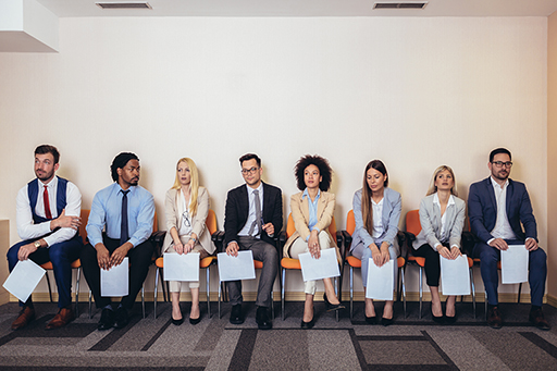 A photograph of 8 people sat on chairs in a line against a wall, each with papers in their hands.