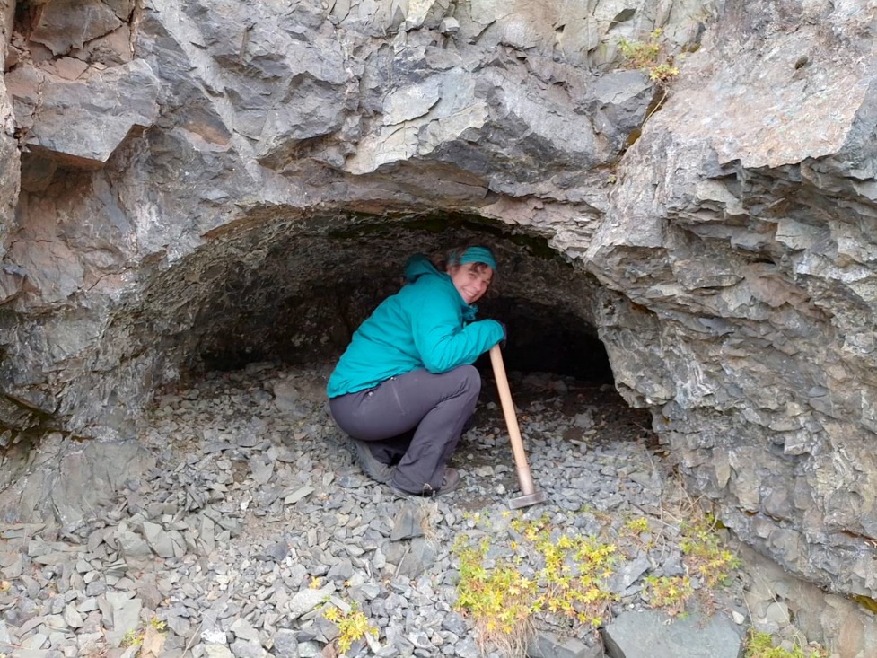 Julia, crouching in a cave cut into a grey rock, wearing a turquoise jacket and hat and grey trousers, carrying a hammer.