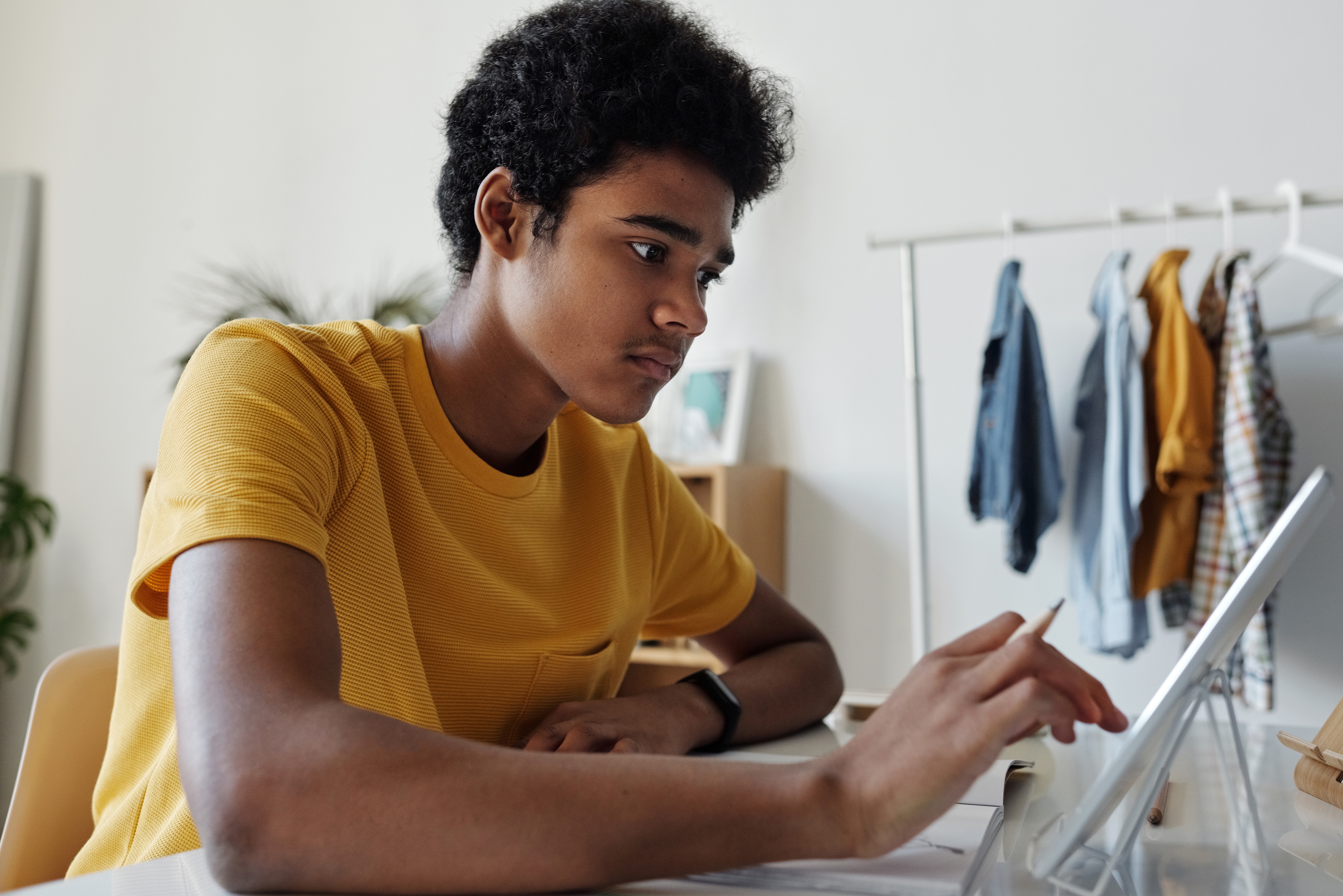 A young man wearing a yellow T-shirt, sitting in front of a laptop and looking at the screen