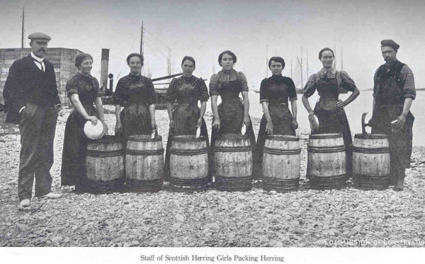Six herring fisherwomen standing in a row behind barrels, flanked at each end of the row by a man.