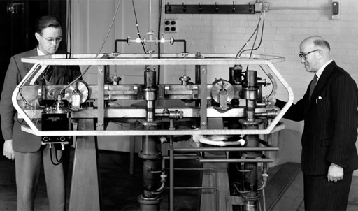 This is a black-and-white photograph of two men examining the first practical atomic clock.