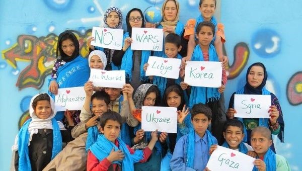 A group of children, some wearing hijabs, holding signs say no wars and names of various countries.
