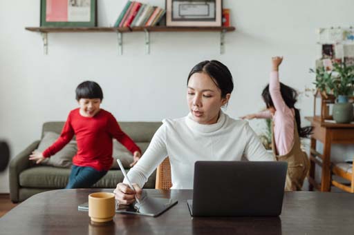 A person sitting at a table with a laptop, with two young children in the background.