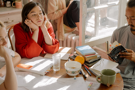 Two people at a table covered with books and notepads. One is resting their head on their hands and the other is reading a book.