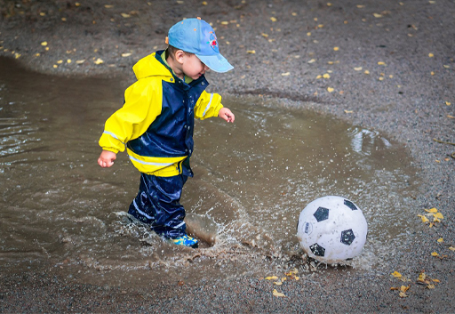 A child kicking a ball through a puddle.