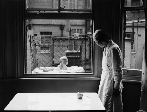 A black-and-white photograph of a baby in a cage next to a window, with the mother looking on.