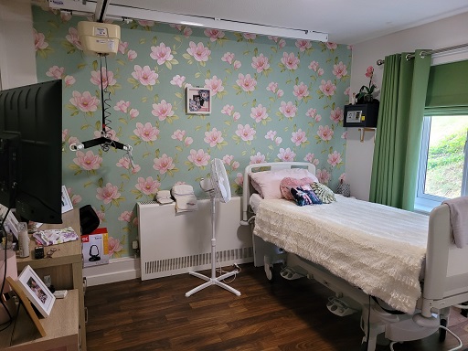 A photograph of Susie’s bedroom. There is green and pink floral wallpaper on one wall. With an adjustable height bed, hoist on ceiling tracking and circulating fan standing on the floor beside the bed.