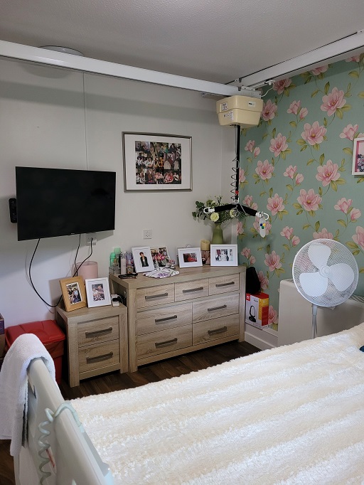 Photograph of Susie’s bedroom. Two chests of drawers displaying photographs of Susie and her family, and a wall mounted television