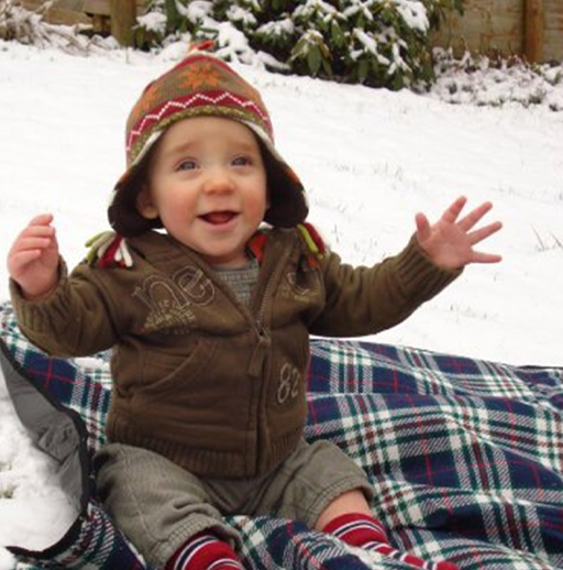 A toddler sitting on a blanket on a snowy lawn.