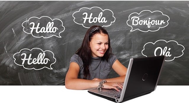 A girl sitting looking at a laptop, in front of a blackboard with hello written in different languages