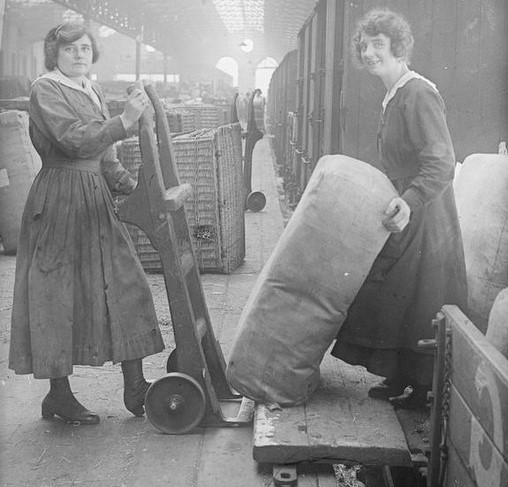 Two ladies moving cargo at the rail station during the first world war. Looking at the camera, smiling.