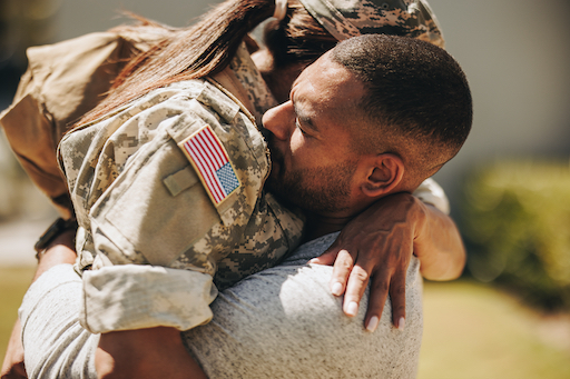 Close-up head-and-shoulders photograph of a man hugging a woman. The man wears a grey long-sleeved T-shirt. The woman’s face is not visible but she has a long ponytail and wears a military camouflage uniform with a US flag stitched to the pocket at the top of her arm.