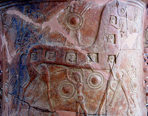 A close-up image of a piece of pottery, decorated in relief with a stylised image of a horse. The horse has several square ‘windows’ along its body and neck, and in each of the windows is a face. Surrounding the horse are several figures carrying shields and weapons, and wearing helmets. The pottery is terracotta in colour, with several cracks. Also visible are the remnants of a coat of dark blue paint, which has mostly worn away.