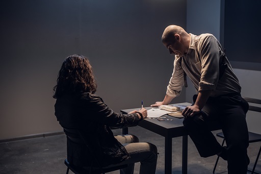 A woman sits at a desk in a darkened interview room. A man sits on the table in front of her. They are both looking at a document she is signing.