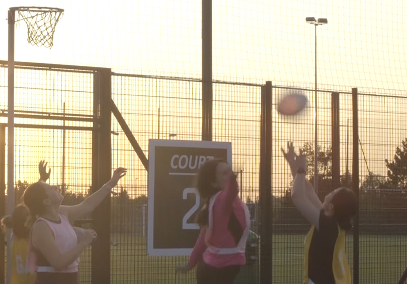 A photograph of people playing netball on a netball court. Someone is taking a shot in front of the netball hoop.