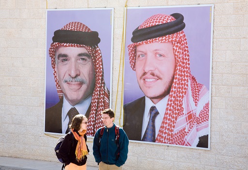 This image shows two people standing in front of two large posters mounted on the external wall of a building. The posters both depict men wearing suits and ties with red-and-white-checked headscarves held in place with black bands. The man on the left is older than the man on the right. Both wear small beards.