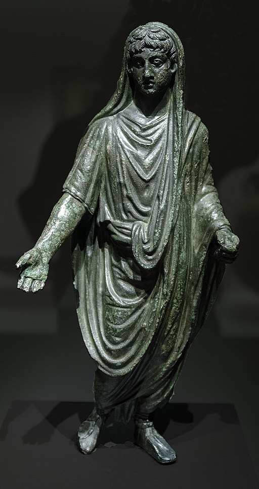 This image shows a bronze statuette of a man standing with his arms stretched out somewhat from his sides. He is holding something in his left hand. He wears a loose, short-sleeved tunic and an imperial toga. In this image, the top diagonal of the toga has been pulled up over the back of the man’s head. He has short, curly hair.