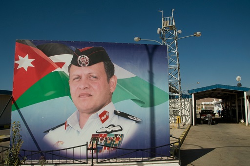 This image shows a large poster mounted next to the entrance to a military camp, consisting of an entranceway and an observation tower. The poster depicts the head, neck, shoulders and chest of a middle-aged man with short hair in front of the Jordanian national flag. The man is wearing military uniform consisting of a cap with a badge and a shirt displaying a multitude of medals and honours on the left chest and shoulders. He is clean-shaven.
