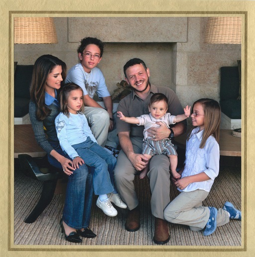This image shows a photograph of a family in a living room. The family consists of: a young-middle-aged woman with long, dark hair in jeans, a shirt and jumper; a middle-aged man with short hair and a short beard wearing a brown shirt and beige trousers; on the woman’s lap, a girl of about 5-6 years wearing jeans and a jumper, on the man’s lap, a baby of about 12 months, kneeling next to the man, a girl of about 10-12 years wearing a shirt and trousers, seated behind the couple a boy of about 10 years in a T-shirt and trousers.
