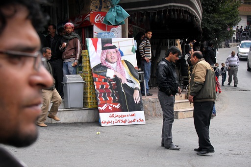 This image shows a large poster mounted in a covered market area. There are several people walking around and talking. The poster depicts a seated middle-aged man with his right arm resting on a pile of folded Arabic-style carpets. The man is wearing a long, sleeved black robe and a red-and-white-checked scarf over his head, held in place with a black band. He wears a short beard.