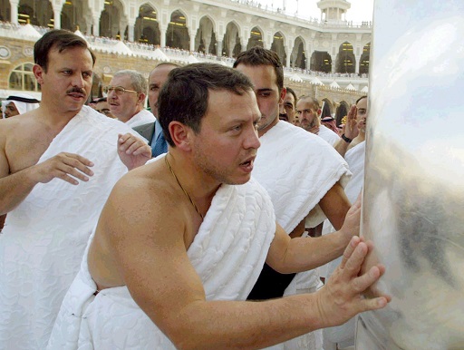 This image shows the top half of a clean-shaven middle-aged man surrounded by other men in an enclosed courtyard. He is wearing nothing but a large white cloth draped around his body and over his left shoulder. He is leaning forward to touch a large object in front of him with both hands, and is speaking.