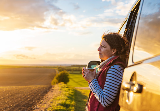 A person holding a hot drink standing against a van, looking into the distance.