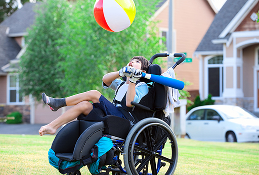 A child in a wheelchair holding a baseball bat and aiming for a beach ball above
