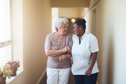 An elderly woman walking arm in arm with a care worker down a corridor whilst both smiling